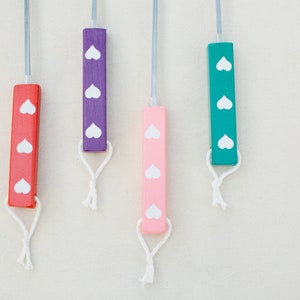 May include: Five wooden skewers with white heart designs. The skewers are painted in different colors: red, purple, pink, and teal.