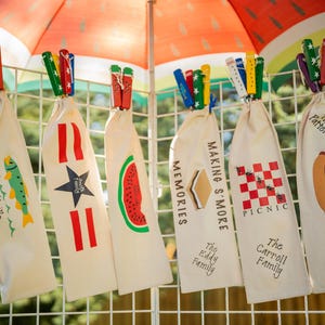 May include: A display of personalized canvas bags, each clipped to a wire grid with colorful clothespins. The bags feature various designs, including a fish, star, watermelon, s'more, picnic, and hot dog, with family names and text.
