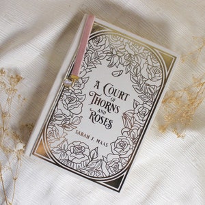 May include: A white book with a gold and black floral design on the cover. The title of the book is "A Court of Thorns and Roses" by Sarah J. Maas.
