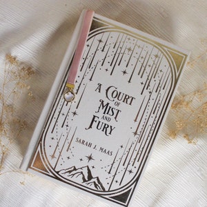 May include: A white book with a gold design on the cover. The title is "A Court of Mist and Fury" by Sarah J. Maas. The book is on a white surface with dried flowers.