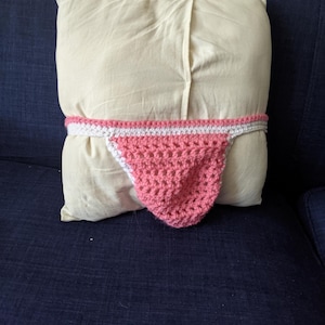 May include: A cream-colored pillow with a pink and white crocheted thong. The thong is wrapped around the pillow, with the pink portion hanging down. The pillow is on a dark blue surface.