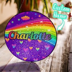 May include: Round, rainbow-themed suncatcher with the name "Charlotte" in colorful letters. The design includes a rainbow, stars, clouds, and hearts on a purple background. A chain is attached for hanging.