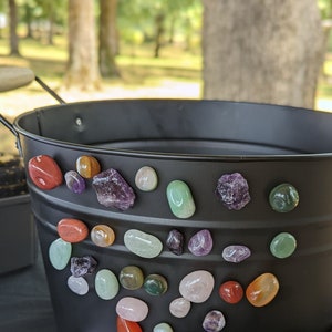 May include: A black metal planter pot with a variety of colorful stones and crystals glued to the rim. The stones are in various shapes and sizes, including round, oval, and irregular.
