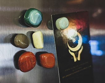 May include: A collection of seven smooth, polished stones in various colors, including green, brown, white, and red, are arranged on a silver surface. The stones are displayed next to a black card with a gold crescent moon and hands design.
