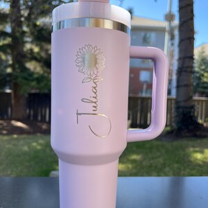 May include: A light purple insulated tumbler with a handle, a white lid, and a silver band. The tumbler features a gold sunflower design and the name "Julian" in a script font. The tumbler is designed for hot or cold beverages.
