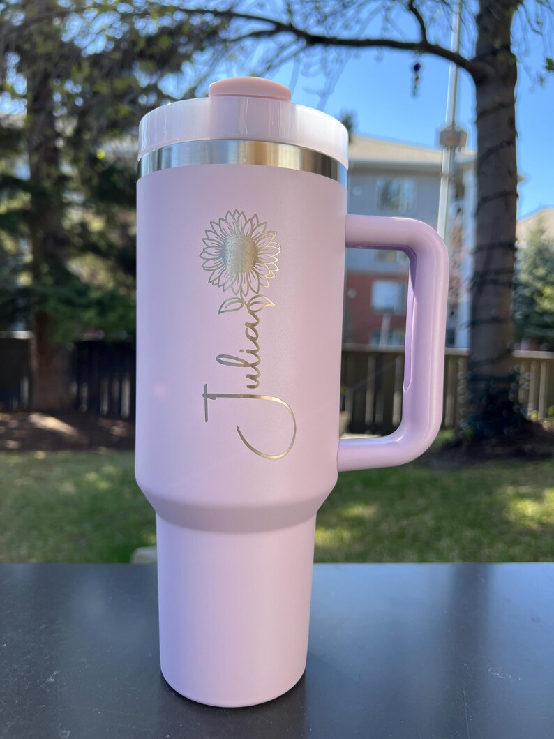 May include: A light purple insulated tumbler with a handle, a white lid, and a silver band. The tumbler features a gold sunflower design and the name "Julian" in a script font. The tumbler is designed for hot or cold beverages.