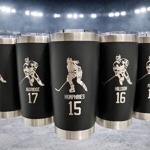 Op de afbeelding: Zwarte ge&iuml;soleerde bekers met zilveren accenten, elk met een hockey speler afbeelding en een naam en nummer. Deze bekers zijn ideaal voor sportfans en voor warme of koude dranken.