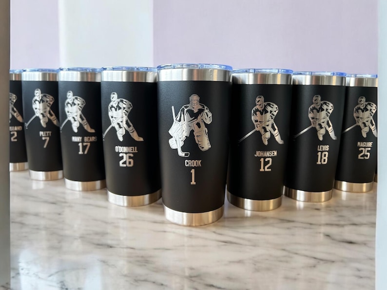 Op de afbeelding: Zwarte ge&iuml;soleerde bekers met zilveren deksels en bodems. Elke beker heeft een zilveren hockey speler afbeelding en een naam of nummer. De bekers zijn gepersonaliseerd met namen en nummers.