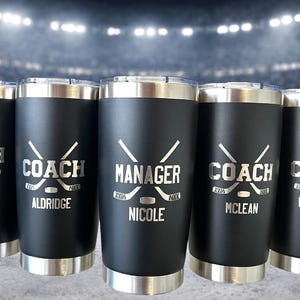 Op de afbeelding: Vijf zwarte ge&iuml;soleerde bekers met zilveren randen en deksels. Elke beker heeft een hockey-thema ontwerp met het woord "COACH" of "MANAGER" en een naam. De achtergrond toont een ijshockeybaan.