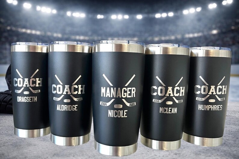 Op de afbeelding: Vijf zwarte ge&iuml;soleerde bekers met zilveren randen en deksels. Elke beker heeft een hockey-thema ontwerp met het woord "COACH" of "MANAGER" en een naam. De achtergrond toont een ijshockeybaan.