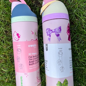 May include: Two Owala water bottles with labels. The left bottle is light pink with Hello Kitty graphics, a blue and mint lid, and a pink carry loop. The right bottle is light pink with purple and green bow graphics, a cream and purple lid.