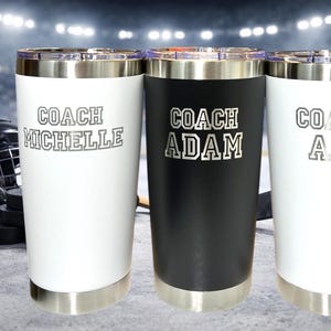 Op de afbeelding: Drie roestvrijstalen ge&iuml;soleerde bekers, twee witte en &eacute;&eacute;n zwarte, met zilveren randen en bodems. Elke beker heeft een naam en het woord "COACH" erop gedrukt. Hockeyuitrusting is op de achtergrond.