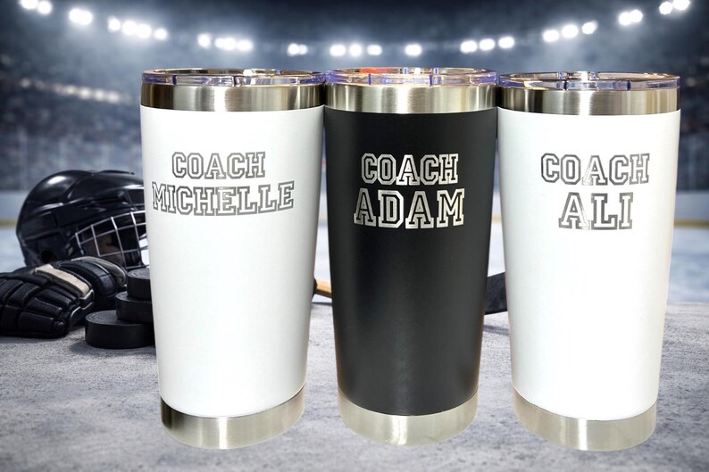 Op de afbeelding: Drie roestvrijstalen ge&iuml;soleerde bekers, twee witte en &eacute;&eacute;n zwarte, met zilveren randen en bodems. Elke beker heeft een naam en het woord "COACH" erop gedrukt. Hockeyuitrusting is op de achtergrond.