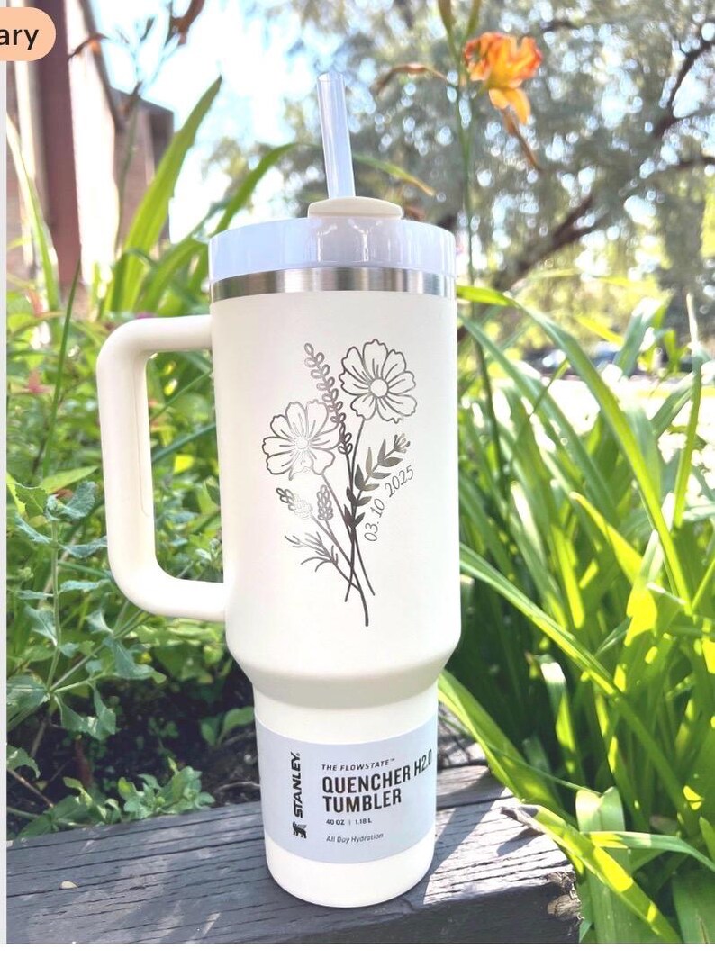 May include: Cream-colored Stanley Quencher H2.0 tumbler with a white handle and lid, and a clear straw. The tumbler features a floral design with the date 03.10.2025. The tumbler is 40 oz / 1.18 L and is designed for all-day hydration.