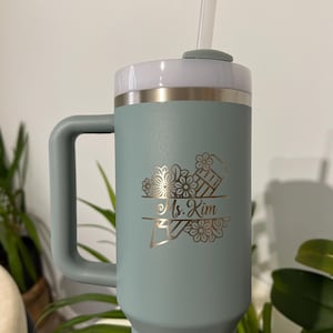 May include: A teal insulated tumbler with a handle, white lid, and straw. The tumbler features a gold floral design with the name "Ms. Kim" engraved. The tumbler is designed to keep drinks cold for hours.
