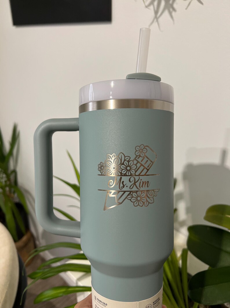May include: A teal insulated tumbler with a handle, white lid, and straw. The tumbler features a gold floral design with the name "Ms. Kim" engraved. The tumbler is designed to keep drinks cold for hours.