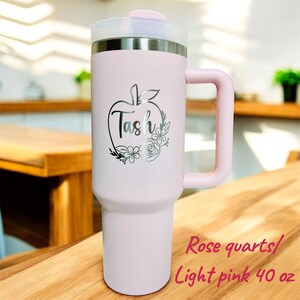 May include: A light pink 40 oz tumbler with a handle and a clear lid. The tumbler has a silver apple design with the name "Tash" and floral accents. The text "Rose quartz/ Light pink 40 oz" is also visible.