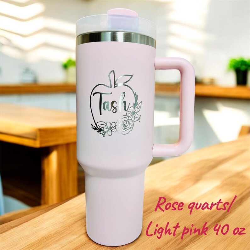 May include: A light pink 40 oz tumbler with a handle and a clear lid. The tumbler has a silver apple design with the name "Tash" and floral accents. The text "Rose quartz/ Light pink 40 oz" is also visible.