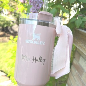 May include: A light pink Stanley tumbler with a handle and a clear straw. The tumbler has the Stanley logo and the name "Mrs. Halby" engraved on it. A light pink ribbon is tied around the handle.