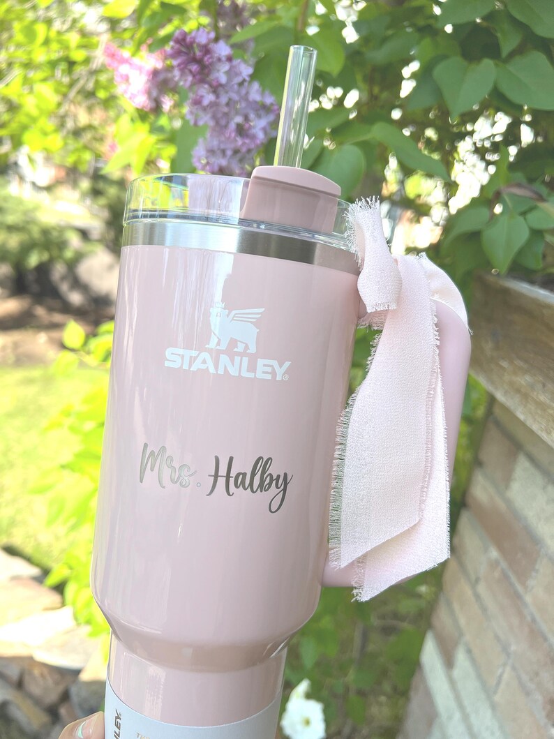 May include: A light pink Stanley tumbler with a handle and a clear straw. The tumbler has the Stanley logo and the name "Mrs. Halby" engraved on it. A light pink ribbon is tied around the handle.