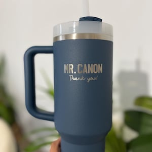 May include: A navy blue insulated tumbler with a handle, a white lid, and a clear straw. The text "MR. CANON Thank you!" is printed on the side. The tumbler is designed to keep beverages cold or hot.