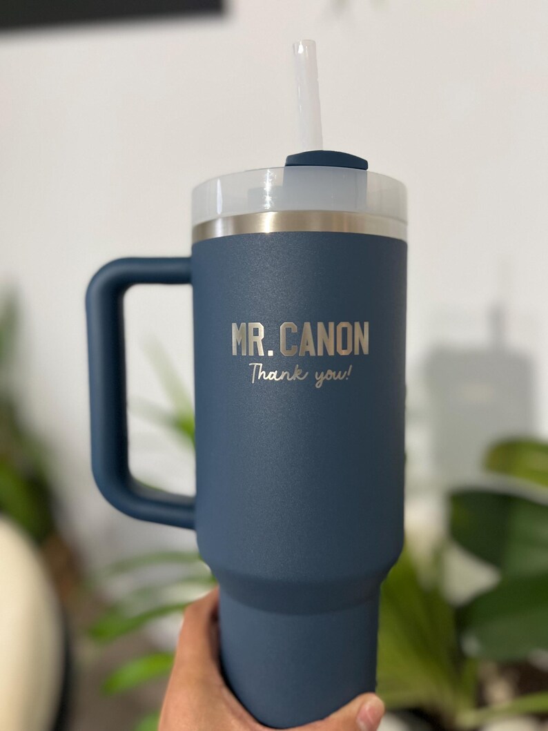 May include: A navy blue insulated tumbler with a handle, a white lid, and a clear straw. The text "MR. CANON Thank you!" is printed on the side. The tumbler is designed to keep beverages cold or hot.