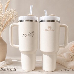 May include: Two cream-colored Stanley tumblers with handles and straws. One tumbler has the word "Bride" in script, the other has the Stanley logo. Both have silver accents and white lids. The tumblers are on a white surface.