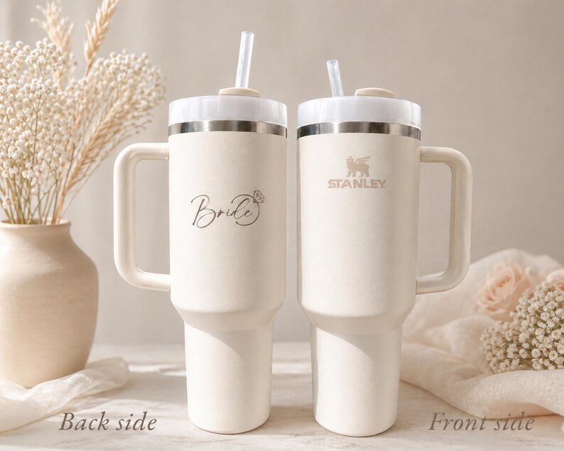 May include: Two cream-colored Stanley tumblers with handles and straws. One tumbler has the word "Bride" in script, the other has the Stanley logo. Both have silver accents and white lids. The tumblers are on a white surface.