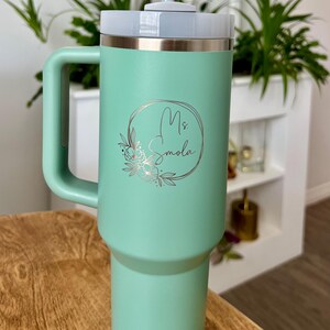May include: A mint green insulated tumbler with a handle, a white lid, and a silver rim. The tumbler features the name "Ms. Smola" in a floral design. A reusable straw is inserted into the lid. The tumbler is on a wooden surface.