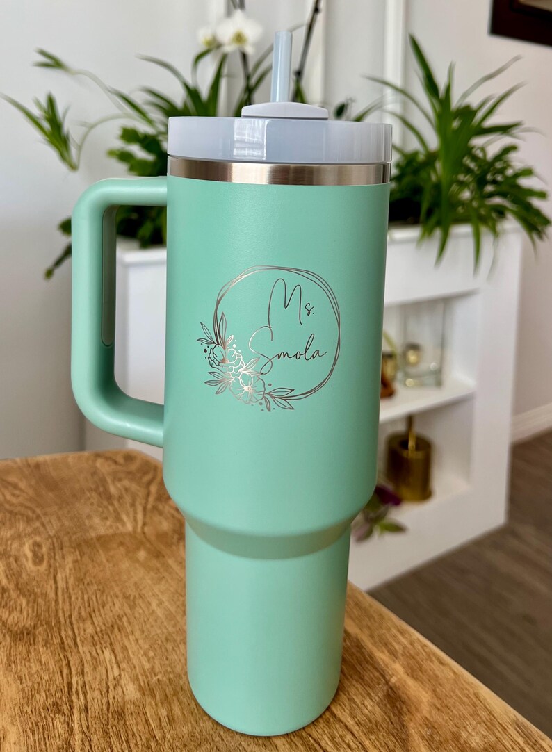 May include: A mint green insulated tumbler with a handle, a white lid, and a silver rim. The tumbler features the name "Ms. Smola" in a floral design. A reusable straw is inserted into the lid. The tumbler is on a wooden surface.