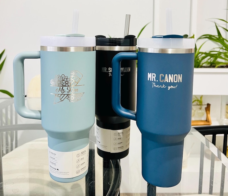 May include: Three insulated tumblers with handles and straws. One is light blue with "Ms. Jim" and floral design, another is black, and the third is blue with "MR. CANON Thank you!" text. All have white lids.