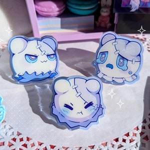 Puede incluir: Tres pines de personajes de dibujos animados azules y blancos con detalles cosidos. Los personajes tienen diferentes expresiones faciales, incluyendo una cara gruñona, una cara triste y una cara sorprendida.