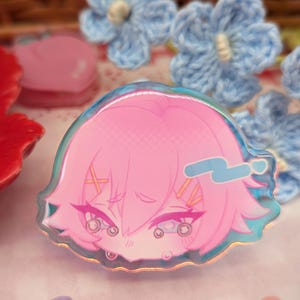 Puede incluir: Una pegatina rosa de personaje de estilo anime con borde holográfico. El personaje tiene pelo rosa, ojos cerrados y un signo de interrogación azul. La pegatina está rodeada de flores azules de ganchillo y otros objetos decorativos.