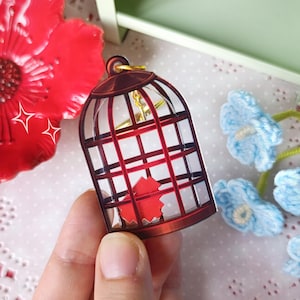 Może przedstawiać: Czerwono-brązowy brelok do kluczy w kształcie klatki dla ptaków ze złotym zapięciem. Brelok ma czerwony wzór w kształcie serca w środku. Duży czerwony kwiat i niebieskie kwiaty szydełkowe w tle.