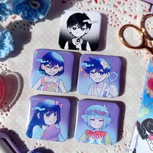 May include: Five square character badges with anime-style illustrations. The badges feature characters with blue hair and various expressions, set against a purple background. One badge has a black and white design. Accessories like scissors and a ribbon are also visible.