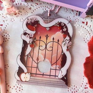 May include: A decorative card featuring a snake entwined around a gate with apples and flowers. The card has a sunset background with a heart-shaped cutout. Pink bunny figurines and a red poppy are in the background.