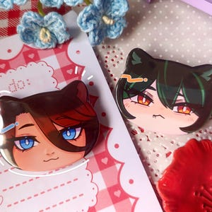 Puede incluir: Dos pines de personajes con forma de gato. Un pin presenta un personaje con cabello castaño y ojos azules, el otro tiene cabello verde y ojos anaranjados. Los pines están sobre una superficie estampada en rosa y blanco con una lista "To do:"