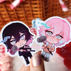 Puede incluir: Dos pegatinas de vinilo troqueladas con personajes de estilo anime que sostienen martillos. El personaje de la izquierda tiene el pelo negro y ropa negra, mientras que el de la derecha tiene el pelo rosa y un suéter beige. Incluyen el texto @nisestarmin.