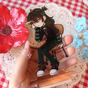 Può includere: Un espositore in acrilico trasparente con un personaggio dei cartoni animati con capelli e vestiti verdi, seduto su una sedia. Il personaggio indossa una camicia nera a righe verdi, pantaloni verdi e scarpe da ginnastica bianche e nere. L'espositore è tenuto davanti a uno sfondo a scacchi rossi e bianchi.