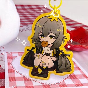 K&ouml;nnte beinhalten: Ein gelber Acryl-Schl&uuml;sselanh&auml;nger mit einem Anime-Charakter mit grauen Haaren, der ein Geb&auml;ck h&auml;lt. Die Figur ist von schwarzen M&uuml;lls&auml;cken umgeben und tr&auml;gt ein schwarzes Outfit. Der Schl&uuml;sselanh&auml;nger hat einen gelben Rand und einen Metallring.