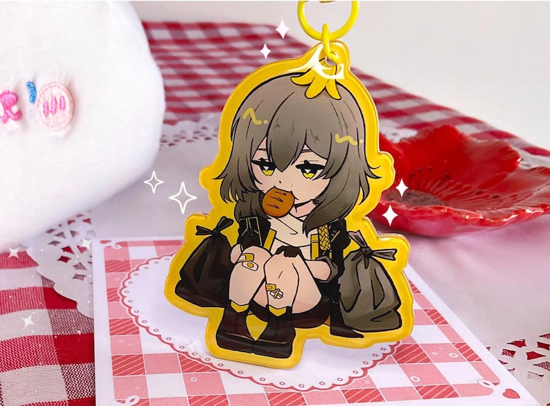K&ouml;nnte beinhalten: Ein gelber Acryl-Schl&uuml;sselanh&auml;nger mit einem Anime-Charakter mit grauen Haaren, der ein Geb&auml;ck h&auml;lt. Die Figur ist von schwarzen M&uuml;lls&auml;cken umgeben und tr&auml;gt ein schwarzes Outfit. Der Schl&uuml;sselanh&auml;nger hat einen gelben Rand und einen Metallring.