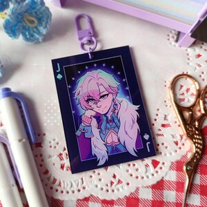 K&ouml;nnte beinhalten: Ein rechteckiger Schl&uuml;sselanh&auml;nger mit einer Charakterillustration, umrahmt von einem dunkelblauen Rand mit einem Pik-Buben-Symbol. Die Figur hat rosa und gr&uuml;ne Haare, ein blaues Outfit und einen wei&szlig;en Pelzkragen. Ein lila Verschluss ist angebracht.