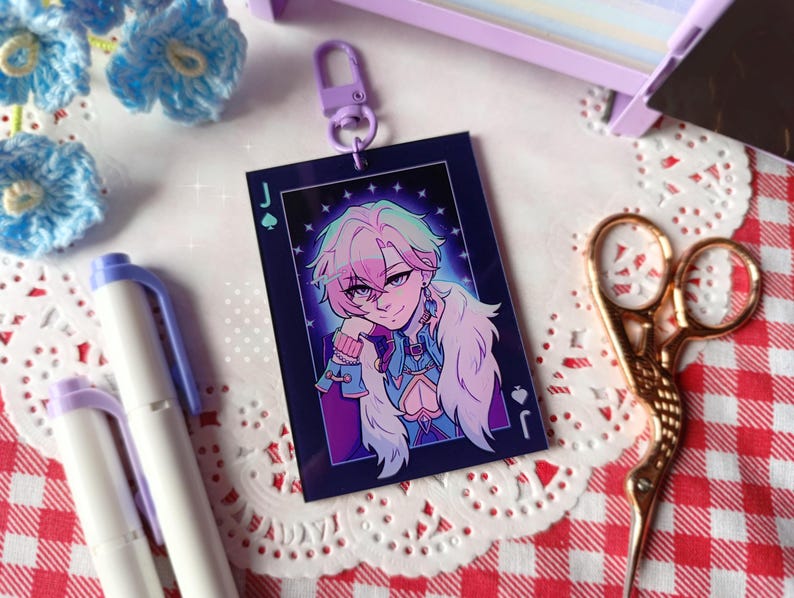 K&ouml;nnte beinhalten: Ein rechteckiger Schl&uuml;sselanh&auml;nger mit einer Charakterillustration, umrahmt von einem dunkelblauen Rand mit einem Pik-Buben-Symbol. Die Figur hat rosa und gr&uuml;ne Haare, ein blaues Outfit und einen wei&szlig;en Pelzkragen. Ein lila Verschluss ist angebracht.