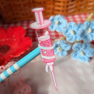 Puede incluir: Un dije de acr&iacute;lico transparente con forma de jeringa llena de l&iacute;quido rosa, con peque&ntilde;os gr&aacute;ficos de corazones y nubes blancas dentro del cilindro. La jeringa tiene un &eacute;mbolo rosa y una tapa de aguja blanca, unida a un bol&iacute;grafo de rayas azules y blancas.