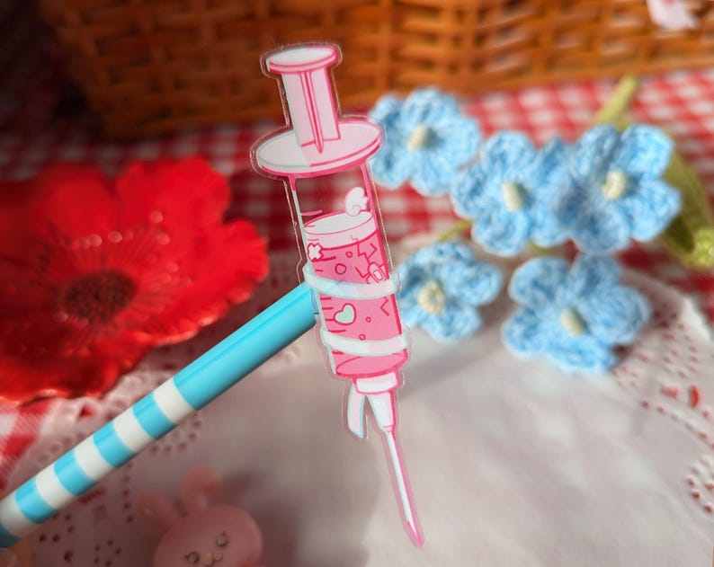 Puede incluir: Un dije de acr&iacute;lico transparente con forma de jeringa llena de l&iacute;quido rosa, con peque&ntilde;os gr&aacute;ficos de corazones y nubes blancas dentro del cilindro. La jeringa tiene un &eacute;mbolo rosa y una tapa de aguja blanca, unida a un bol&iacute;grafo de rayas azules y blancas.