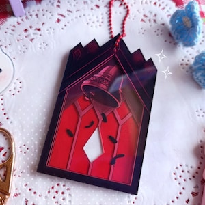 Könnte beinhalten: Ein roter und schwarzer Acryl-Anhänger mit einer Glocke in einem Fenster mit einer Diamantform in der Mitte. Der Anhänger ist an einer silbernen Kette befestigt.