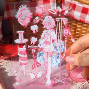 Puede incluir: Una hoja de pegatinas transparente con ilustraciones m&eacute;dicas en rosa y blanco, que incluyen un personaje de enfermer&iacute;a, un soporte para suero, una jeringa, un coraz&oacute;n, dientes, pastillas y alas. El texto @nisestarmin est&aacute; impreso en el centro.