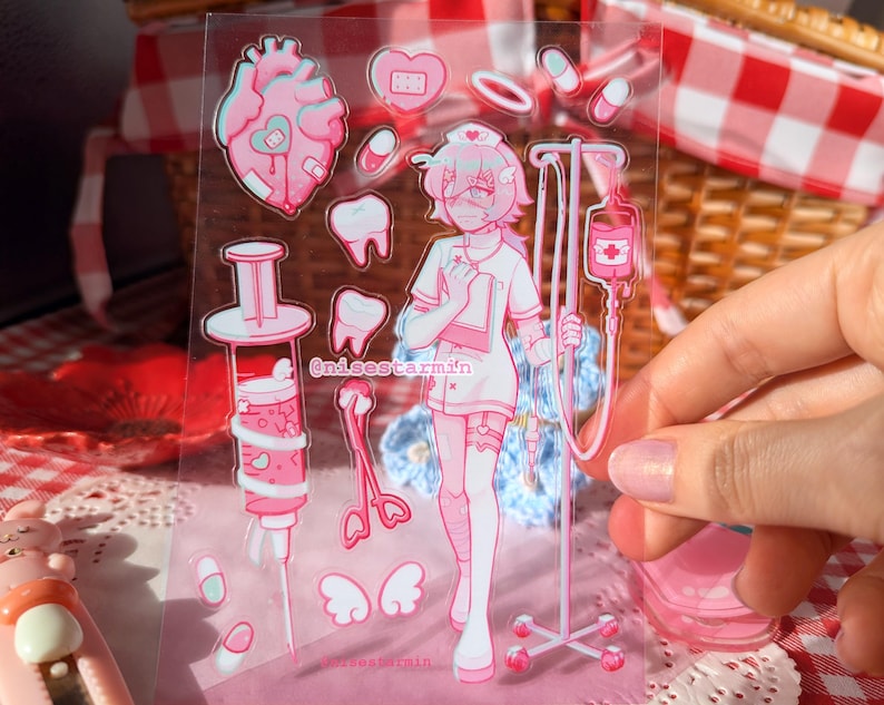 Puede incluir: Una hoja de pegatinas transparente con ilustraciones m&eacute;dicas en rosa y blanco, que incluyen un personaje de enfermer&iacute;a, un soporte para suero, una jeringa, un coraz&oacute;n, dientes, pastillas y alas. El texto @nisestarmin est&aacute; impreso en el centro.