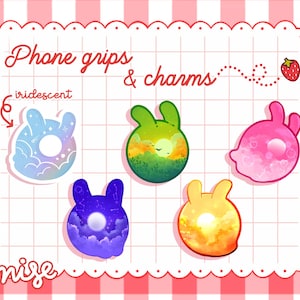 Puede incluir: Cinco agarres para teléfono y dijes con siluetas de conejos con diferentes fondos iridiscentes. Los fondos incluyen un cielo nocturno estrellado, una puesta de sol, una salida del sol, una escena de bosque verde y un cielo rosa con corazones. El texto "Phone grips & charms" está en la parte superior de la imagen. El texto "iridescent" está a la izquierda de la imagen. El texto "nise" está en la parte inferior de la imagen.