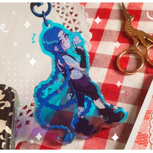 K&ouml;nnte beinhalten: Ein blau-violetter Acryl-Schl&uuml;sselanh&auml;nger mit einer Figur mit langen blauen Z&ouml;pfen. Die Figur tr&auml;gt ein schwarzes Outfit und h&auml;lt einen kleinen Gegenstand. Der Schl&uuml;sselanh&auml;nger ist an einem blauen Metallring befestigt.